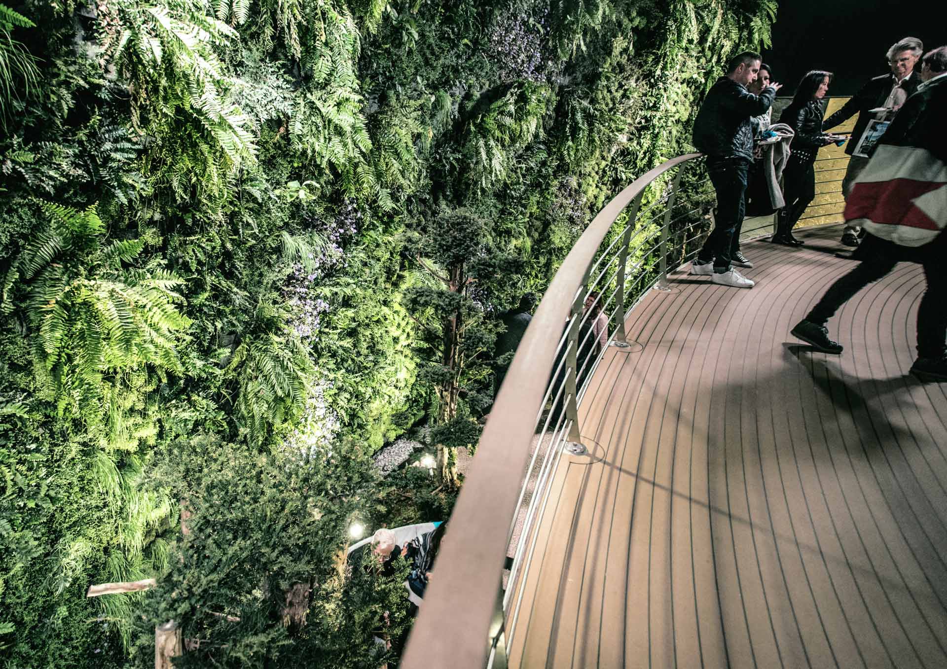 Menschen auf einem geschwungenen Balkon mit Blick auf einen üppigen vertikalen Garten, der nachts Grüne Architektur verkörpert, mit beleuchteten Wegen und stimmungsvoller Beleuchtung.